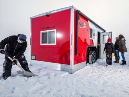 Family standing at the entrance of a bright red Yetti Fish House Angler Edition. The fish house is parked on a lake that is covered in snow.